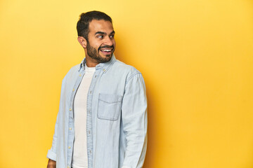 Wall Mural - Casual young Latino man against a vibrant yellow studio background, looks aside smiling, cheerful and pleasant.