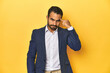 Professional young Latino man in business suit, yellow studio background, pointing temple with finger, thinking, focused on a task.