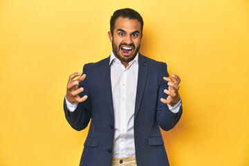 Professional young Latino man in business suit, yellow studio background, screaming with rage.