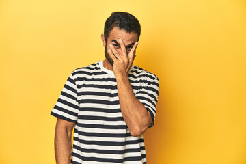 Wall Mural - Casual young Latino man against a vibrant yellow studio background, blink at the camera through fingers, embarrassed covering face.