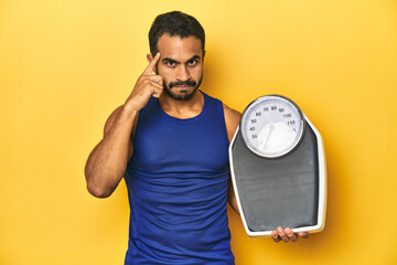 Wall Mural - Fit young Latino man with scale on yellow background, fitness concept, pointing temple with finger, thinking, focused on a task.