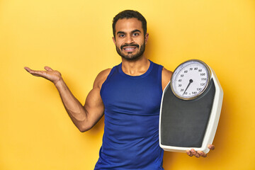 Wall Mural - Fit young Latino man with scale on yellow background, fitness concept, showing a copy space on a palm and holding another hand on waist.