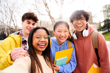 Wall Mural - Group of multi-ethnic high school students taking a selfie outdoors at the university campus holding folders. Looking at camera picture of a diverse colleagues having fun. 