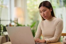 Serious young woman in beige turtleneck working on laptop at home