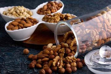 On a gray table, nuts are laid out on a beautiful plate next to a scattered mixture from a glass jar. Assorted nuts in a dried fruit store on a gray textured background