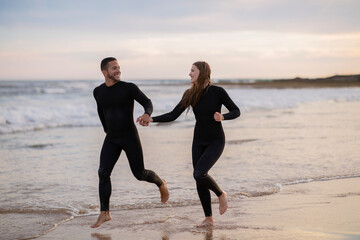 Wall Mural - Cheerful Young Couple Wearing Wetsuits Running On Seashore Together