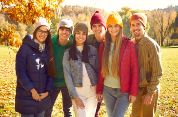 Happy cheerful young friends enjoying fall season together. Group portrait of joyful young people in colorful warm jackets, coats and hats standing in sunny autumn park, looking at camera and smiling