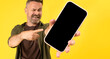 Excited bristle middle-age man holding big smartphone with blank screen in hand, showing close to camera and pointing at device. Big Gadget with empty free space for mock up, banner yellow wall.
