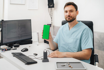 Doctor audiologist holding smartphone and hearing aids that connects to phone for remote communication and adjustments. Telehealth in clinic. Copy space