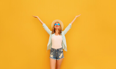 Wall Mural - Happy cheerful caucasian smiling young woman raising her hands up wearing shorts, summer straw hat on yellow background