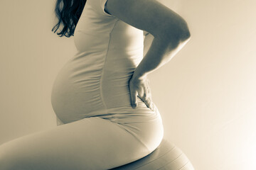 Wall Mural - Pregnant woman with baby bump treats back pain and with the help of a gym ball. Final month of pregnancy - week 36. Side view. White background. Bright shot. Partial toning.