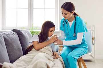 Woman doctor helps sick child take medicine during home visit. Female pediatrician in scrubs sits by sofa with little girl patient, helps her take her medicine, and gives her glass of water to drink