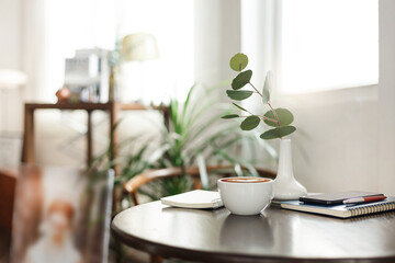 White coffee cup on wooden table and note book, smart phone with ceramic vase and small tree, coffee break after work in cafe with reading book and interior decoration of cafe in the background