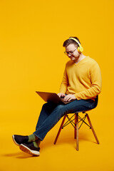 Adult man wearing yellow sweater and eyeglasses, using laptop computer while listening music in headphones, sitting on chair, isolated over orange