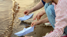 Close Up Of Childrens Hands Play With Paper Boats On Sea Or River In Summer At Sunset. Kids Outdoors By Water