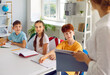 © Studio Romantic - Cute smiling school-age children listen attentively to their teacher in spacious cozy classroom, sitting at desk with pen and school notebook. Side view, from the teacher's back.
