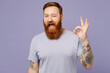 © ViDi Studio - Young cheerful fun redhead bearded man wearing violet t-shirt casual clothes showing okay ok gesture wink blink eye isolated on plain pastel light purple background studio portrait. Lifestyle concept.