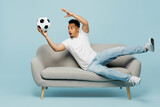 Fototapeta Sport - Full body happy fun cheerful cool young man fan wear t-shirt cheer up support football sport team hold catch soccer ball lay down on grey sofa watch tv live stream isolated on plain blue background.