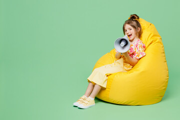 Wall Mural - Full body little child kid girl 6-7 years old wear casual clothes sit in bag chair hold megaphone scream announces discounts sale isolated on plain green background. Mother's Day love family concept.
