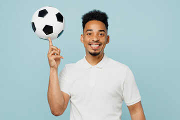 Wall Mural - Young smiling man fan wear basic t-shirt cheer up support football sport team hold in hand soccer ball spin on finger look camera watch tv live stream isolated on plain pastel blue color background.