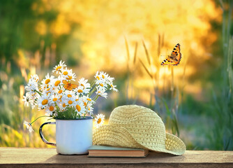 Sticker - chamomile flowers in cup, butterflies, book and braided hat on table in garden, sunny natural abstract background. summer season. Beautiful rustic floral composition. relaxation, harmony atmosphere.