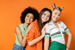 © LIGHTFIELD STUDIOS - Lively multiethnic teenagers with bold makeup wearing stylish casual clothes and hugging blonde girlfriend while standing together on orange background, fashionable and trendy clothes