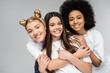 Portrait of cheerful and multiethnic teenage girls in white t-shirts hugging each other and looking at camera while standing isolated on grey, teenage friends having fun together
