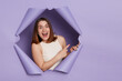 © sementsova321 - Positive delighted excited brunette woman breaking through purple paper hole wearing casual white top pointing away at copy space for promotion advertisement area.