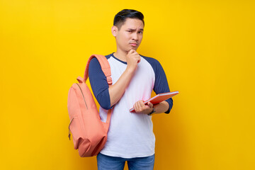 Portrait of pensive young Asian man student wearing t-shirt with backpack holding notebook and thinking about test isolated on yellow background. Education in high school university college concept