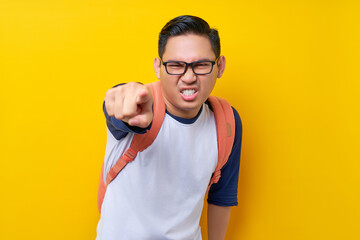 Angry young Asian man student in casual clothes backpack glasses standing pointing finger on camera isolated on yellow background. high school university college concept