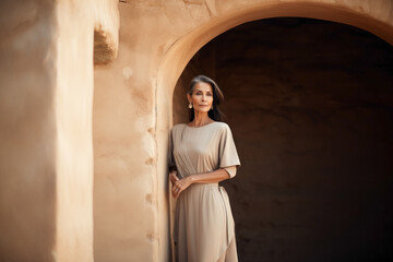 Wall Mural - A beautiful elderly woman model in a linen dress stands near a sandstone wall in the sun.