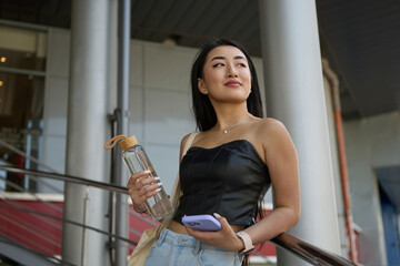 Beautiful Asian female posing outdoor with a glass bottle of water and modern smart phone in hands. Portrait of a cheerful POC woman from Vietnam standing on stairs outdoors