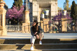 © @skuder_photographer - Beautiful young woman with long brown hair wearing a black and white striped T-shirt and white trousers, sitting on the steps of a square in a park in Seville.