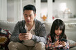 asian father and daughter sitting on family couch using mobile phone together at home