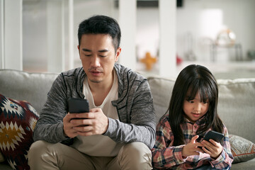 Wall Mural - asian father and daughter sitting on family couch using mobile phone together at home