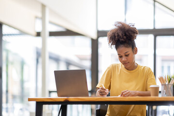 Wall Mural - Business And Education Concept. Smiling african american  sitting at desk working on laptop writing letter in paper documents, free copy space. Happy millennial female studying using laptop	