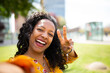 © mimagephotos - Cheerful young african woman taking selfie with peace sign outside