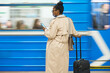 Rear view of young African American female traveler with suitcase and smartphone standing at subway station against mobing blue train