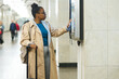 Side view of young African American woman with suitcase scrolling through route map on display while standing at subway station