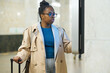 Young serious female traveler in eyeglasses and beige trenchcoat standing in front of display and looking through timetable of subway trains