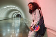 Adolescent guy in stylish casualwear holding skateboard while standing by wall in long underground tunnel of subway station and waiting for train