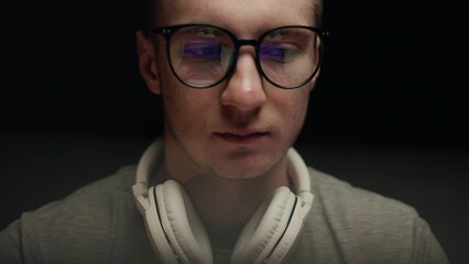 Concentrated young professional programmer in computer glasses with monitors reflection works online late. Serious hacker, IT specialist using laptop computer. Esport player in virtual competition.