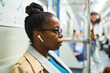 Side view of young serious African American woman in eyeglasses and casualwear listening to music in earphones while traveling by metro