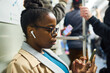 Young serious African American businesswoman in earphones using mobile phone while sitting in subway train and riding home