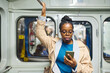 Young serious African American woman in eyeglasses holding by handrail while riding in subway train and watching video in smartphone