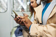 Hands of young African American woman in stylish beige trenchcoat scrolling in mobile phone while sitting in subway train and riding to work
