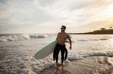 Wall Mural - Surfer Guy. Young Man In Wetsuit Carrying Surfboard While Walking On Seashore