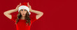 Young caucasian woman with curly hair wearing christmas hat standing on red background making fish face with lips, crazy and comical gesture. Funny expression.