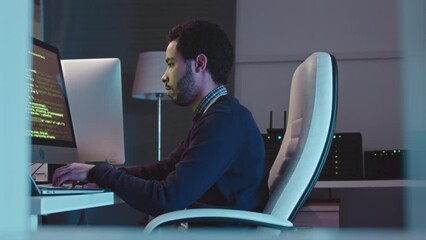 Side view medium shot of Middle Eastern male system administrator program coding on desktop computer in dark office