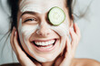 Happy and smiling young woman with facial mask and slice of fresh cucumber on her face and eyes